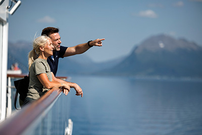 Ein Paar steht an Deck eines Havila Schiffs, blickt auf einen Fjord in Norwegen. Der Mann zeigt mit dem Finger auf die Berge. Ein Paar steht an Deck eines Havila Schiffs, blickt auf einen Fjord in Norwegen. Der Mann zeigt mit dem Finger auf die Berge.