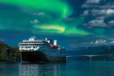 Havila Kystruten Schiff fährt unter leuchtenden grünen Nordlichtern (Aurora Borealis) und einem dramatischen Himmel entlang der norwegischen Küste. Havila Kystruten Schiff fährt unter leuchtenden grünen Nordlichtern (Aurora Borealis) und einem dramatischen Himmel entlang der norwegischen Küste.