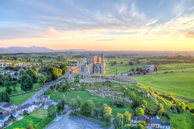 Mittelalterliche Klosterruine Rock of Cashel auf einem grünen Hügel in Irland vor einer weiten Landschaft bei Sonnenuntergang und blauem Himmel.