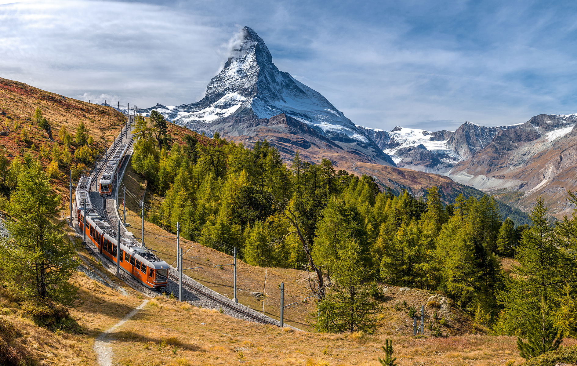 Eine orangefarbene Zahnradbahn der Gornergrat Bahn fährt auf einem steilen, kurvigen Hang in den Schweizer Alpen. Im Hintergrund erhebt sich das markante Matterhorn mit seiner schneebedeckten Spitze unter einem dramatischen Himmel mit Wolken und blauem Himmel. Die Landschaft ist von grünen Nadelbäumen und herbstlich gefärbten Gräsern geprägt, die den Gebirgshang bedecken.